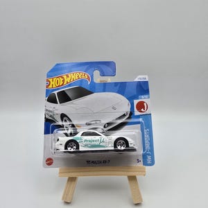 Puede incluir: Un coche blanco '95 Mazda RX-7 Hot Wheels. El coche presenta el texto "Project U" y se muestra en una tarjeta azul y blanca. El embalaje incluye el logotipo de Hot Wheels y el texto "HW J-IMPORTS".