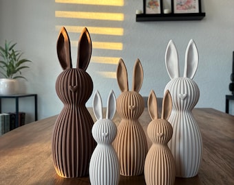 Può includere: Una collezione di cinque figurine decorative a forma di coniglio in varie dimensioni e colori, tra cui marrone, beige e bianco. I coniglietti hanno un design a coste e lunghe orecchie, disposti su un tavolo di legno. Sfondo con arte floreale incorniciata.