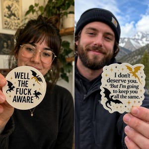 Pode incluir: Duas pessoas segurando adesivos com texto e ilustrações de dragões. Um adesivo diz "WELL THE FUCK AWARE" com dragões pretos e dourados. O outro adesivo diz "I don't deserve you. But I'm going to keep you all the same." com dragões dourados e pretos.
