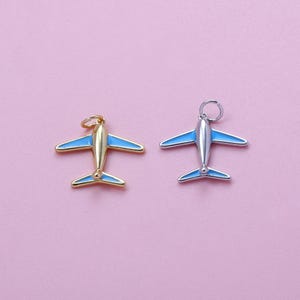 May include: Two airplane charms with blue wings and a gold and silver finish. The charms have a small loop at the top for attaching to a necklace or bracelet. The background is a solid pink color.