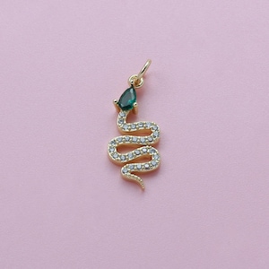 Op de afbeelding: Een goudkleurige slangenhanger met een druppelvormige groene edelsteen kop en een lichaam bezet met kleine, heldere kristallen. De hanger is ontworpen met een lus aan de bovenkant om op te hangen en staat tegen een zachtroze achtergrond.