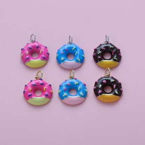 May include: Six colorful donut charms with silver-tone loops for jewelry making. The donuts feature pink, blue, and chocolate frosting with sprinkles. Each donut has a yellow or pink base and a light pink or yellow bottom.