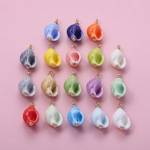 May include: A collection of colorful ceramic seashell charms in various shades, including blue, purple, green, and yellow. Each shell has a gold-colored loop at the top for attaching to jewelry or other crafts. The background is a soft pink.