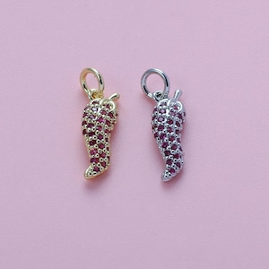 Puede incluir: Dos colgantes con forma de chile. Uno es dorado, el otro plateado. Ambos están incrustados con pequeñas piedras preciosas rojas. Los colgantes tienen un lazo en la parte superior para sujetarlos a una cadena o pulsera. El fondo es rosa claro liso.