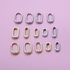 May include: An assortment of oval-shaped metal clasps in gold and silver tones. The clasps are arranged on a pink background, showcasing various sizes. These jewelry findings are suitable for crafting and accessories.