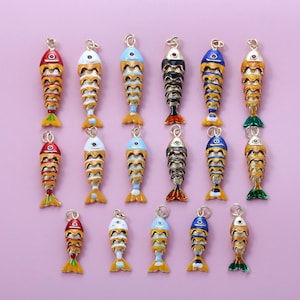 May include: A selection of decorative fish charms in various colours, including red, yellow, blue, and black, with gold detailing. The charms are arranged on a light purple background. Each charm has a unique design and is suitable for jewellery making.