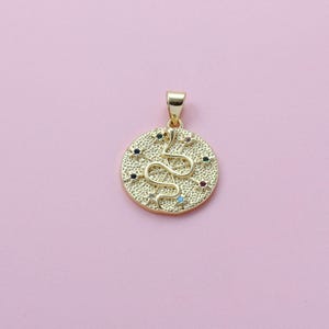 May include: Gold-colored pendant with a snake design. The round pendant features a textured surface with a snake in the center and small, colorful gemstones. The pendant has a loop at the top for a chain, set against a pink background.
