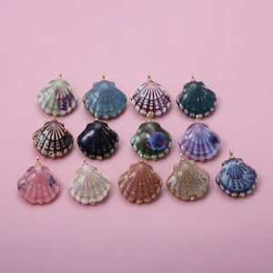 May include: A collection of ceramic seashell pendants in various colors, including blue, green, pink, and brown. Each shell has a gold-colored loop for hanging. The pendants are arranged on a pink background.