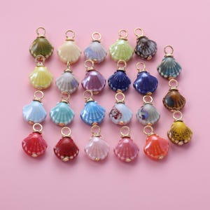 以下が含まれることがあります： 緑、黄色、青、赤、紫など、さまざまな色の貝殻チャームのコレクション。各チャームには、ジュエリーやその他の工芸品に取り付けるための金色のループが付いています。背景は無地のピンク色です。