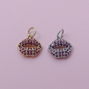 May include: Two lip-shaped pendants. One is gold-toned with red gemstones, the other is silver-toned with red gemstones. Both have a loop at the top for hanging. The background is a solid pink.
