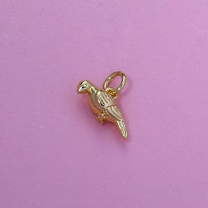 May include: A gold-colored bird charm with a small loop for attaching to a necklace or bracelet. The bird is detailed with etched feathers and a curved beak. The charm is set against a pink background.