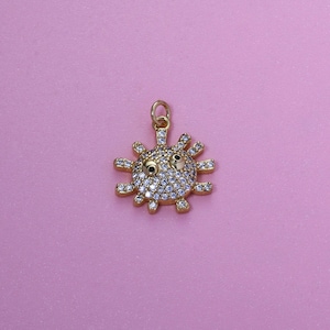 Puede incluir: Un colgante de sol dorado con una cara sonriente, incrustado con pequeños cristales transparentes. El colgante tiene un lazo en la parte superior para una cadena y está colocado sobre un fondo rosa. El sol tiene rayos puntiagudos que se extienden hacia afuera.