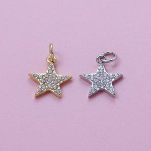 May include: Two star-shaped pendants. One is gold-toned, the other silver-toned. Both pendants are encrusted with small, clear gemstones. Each pendant has a small loop at the top for attaching to a chain or bracelet. The background is a solid pink color.