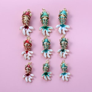 May include: Nine colorful fish-shaped charms with gold-colored accents. The charms feature red, green, and white enamel details. The fish have detailed scales and fins, and are arranged on a pink background. These charms are suitable for jewelry making.