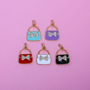 以下が含まれることがあります： ゴールドトーンのハンドルとリボンが付いた、5つのカラフルなハンドバッグチャーム。 ターコイズ、パープル、レッド、ホワイト、ブラックのハンドバッグ。 各チャームには、ゴールドトーンのハンドルとラインストーンのデザインのリボンが付いています。