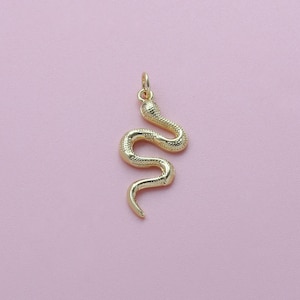 May include: A gold-colored snake pendant with a textured body and a loop at the top for a chain. The snake is in a curved, winding pose. The background is a solid light pink.