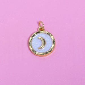 Puede incluir: Colgante dorado con diseño de media luna. El colgante presenta un fondo de esmalte blanco y un marco dorado. La luna es dorada y está sobre el esmalte blanco. El colgante está sobre un fondo rosa.