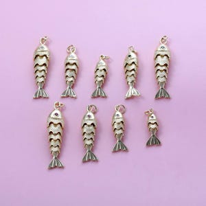 Può includere: Ciondoli a forma di pesce dorato con squame e pinne caudali strutturate su sfondo rosa. Ogni ciondolo ha un anello per il fissaggio ai gioielli. I ciondoli variano di dimensioni, alcuni con piccole gemme.