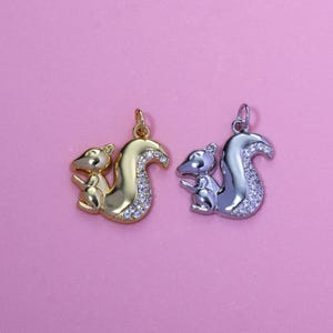 May include: Two squirrel pendants, one gold-toned and one silver-toned, each with a curved tail embellished with small, clear crystals. The pendants are displayed against a pink background, showcasing their detailed design and craftsmanship.