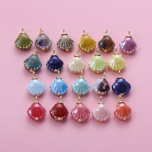 Könnte beinhalten: Eine Sammlung von Muschel-Anhängern aus Keramik in verschiedenen Farben, darunter Grün, Blau, Gelb und Rosa. Jeder Anhänger hat eine goldfarbene Schlaufe zum Befestigen an einer Kette oder einem anderen Schmuckbestandteil. Die Anhänger sind auf einem rosa Hintergrund angeordnet.