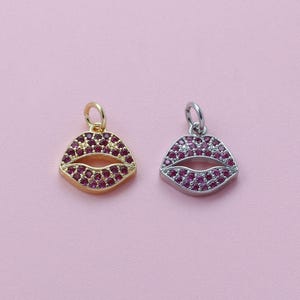 May include: Two lip-shaped pendants. One is gold-toned, the other silver-toned. Both are encrusted with small, round, ruby-colored stones. The pendants have a bail at the top for attaching to a chain or bracelet. The background is a solid, light pink.