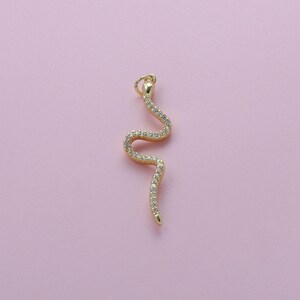 May include: A gold-colored snake pendant with a curved design. The pendant is encrusted with small, clear gemstones. The snake's head has a small loop for attaching to a chain. The background is a solid light pink color.