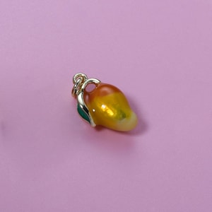 May include: A mango-shaped charm with a gold-colored bail and a green leaf. The mango is painted with a gradient of yellow, orange, and red. The charm is set against a pink background.