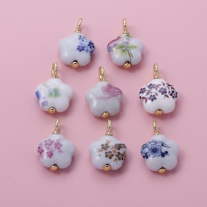 Puede incluir: Nueve colgantes de porcelana blanca en forma de estrella con lazos y detalles dorados. Cada colgante presenta un diseño floral diferente en tonos azules, morados, rosas y marrones. Los colgantes están dispuestos sobre un fondo rosa claro.