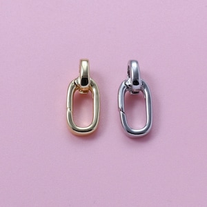 May include: Two metal oval-shaped charms, one gold-toned and one silver-toned, are displayed against a pink background. Each charm has a loop at the top and a hinged clasp. The charms are likely jewelry components or pendants.