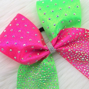 May include: A vibrant cheerleading bow in neon green and hot pink, adorned with sparkling rhinestones. The bow features a central rhinestone-studded band and is set against a fluffy white background. A stylish accessory for cheerleaders.