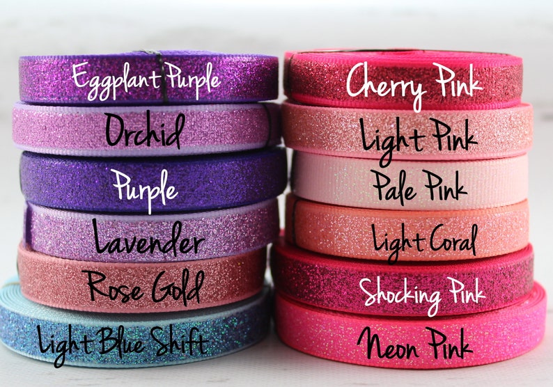 May include: A stack of 12 rolls of glitter ribbon in various shades of purple, pink, and coral. The ribbon is labeled with the following colors: Eggplant Purple, Orchid, Purple, Lavender, Rose Gold, Light Blue Shift, Cherry Pink, Light Pink, Pale Pink, Light Coral, Shocking Pink, and Neon Pink.