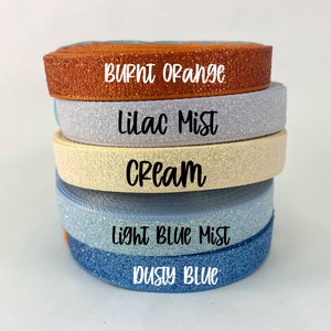 May include: Five rolls of glitter ribbon in different colors. The colors are burnt orange, lilac mist, cream, light blue mist, and dusty blue.