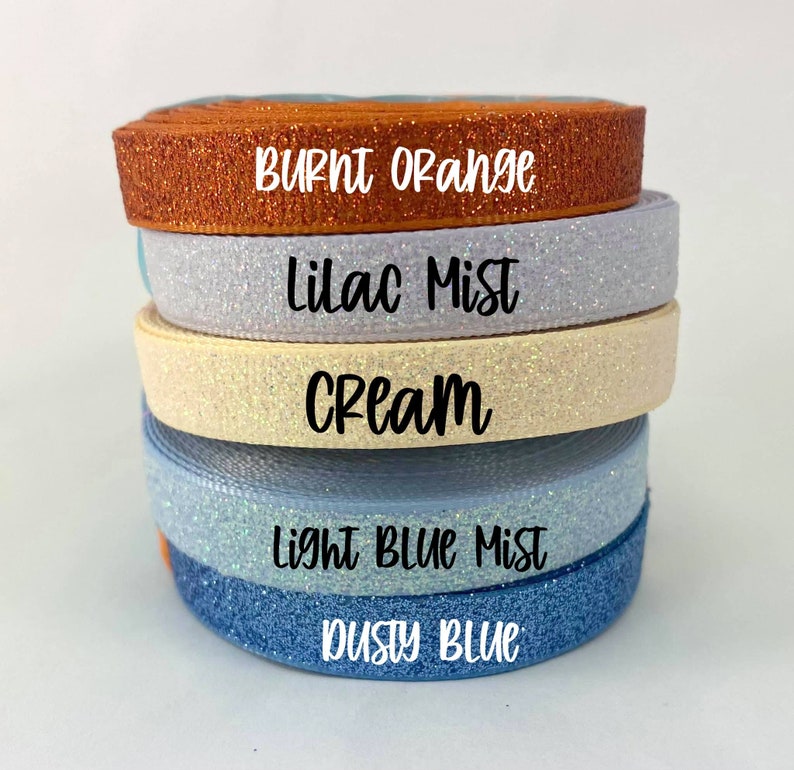 May include: Five rolls of glitter ribbon in different colors. The colors are burnt orange, lilac mist, cream, light blue mist, and dusty blue.