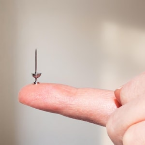 May include: A small, silver-colored metal push pin featuring a sharp, pointed needle-like tip and a rounded, cup-shaped head. The push pin is constructed from a smooth, reflective material and is designed for securing documents or notes to surfaces.