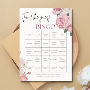 May include: A bridal shower bingo game card with a floral design. The card features a grid with various prompts like "Loves coffee" and "Has a tattoo." The text "Find the guest" and "BINGO" are at the top. The card is on a wooden tray.