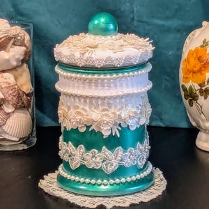 May include: Teal decorative jar with a white lid and white lace trim. The jar is decorated with white lace, pearls, and rhinestones.