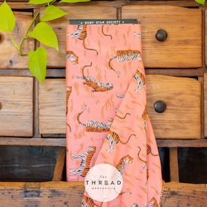 May include: A roll of pink fabric with a repeating pattern of orange and black tigers. The fabric is labeled "Ruby Star Society" and "The Thread Mercantile". The fabric is displayed on a wooden dresser.