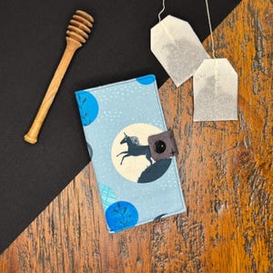 May include: A light blue fabric journal cover with a unicorn design, a brown leather strap, and a black button closure. A wooden honey dipper and two tea bags are also visible.