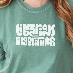 May include: A seafoam green t-shirt with the text "Librarians Not Algorithms" in white, stylized lettering. The shirt has rolled-up sleeves and is paired with blue denim jeans. The overall style is casual and comfortable.