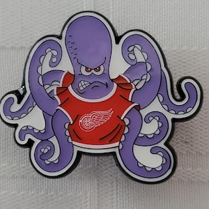 May include: A purple octopus cartoon pin with an angry expression, wearing a red shirt with the Detroit Red Wings logo. The pin has a black outline and white accents.