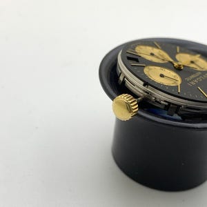 May include: A close-up of a black and gold wristwatch with a black face and gold hands and markers. The watch has three sub-dials and a gold crown. The watch is labeled "BVLGARI AUTOMATIC".
