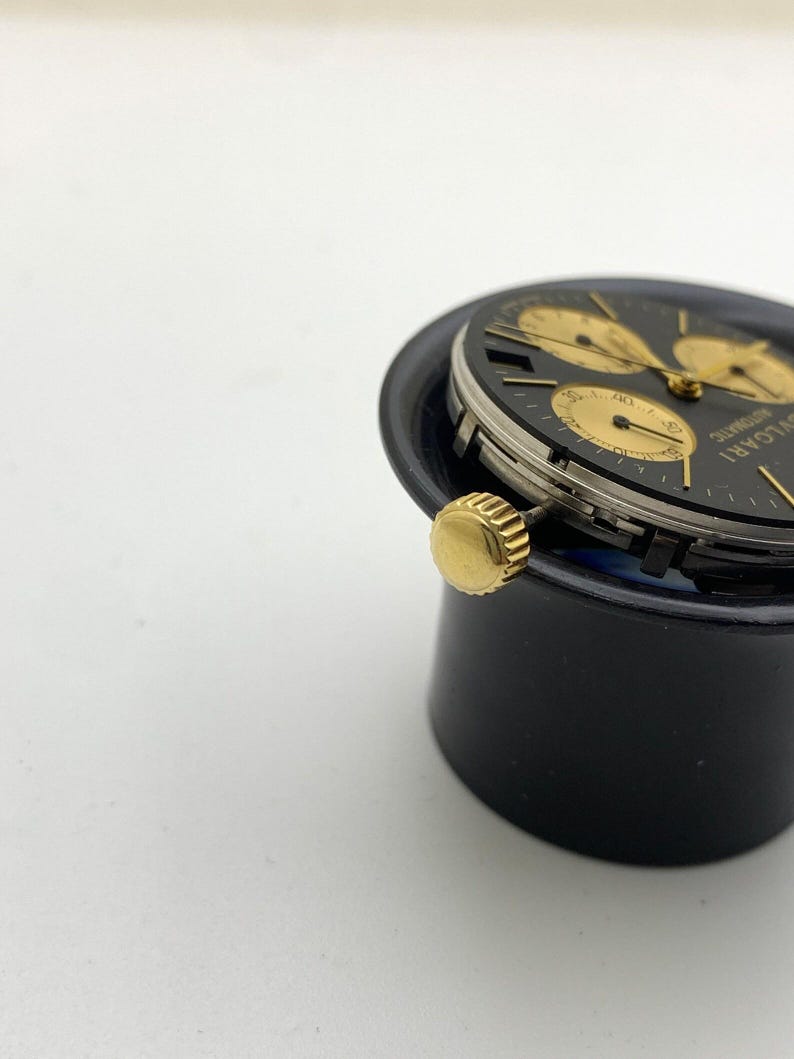 May include: A close-up of a black and gold wristwatch with a black face and gold hands and markers. The watch has three sub-dials and a gold crown. The watch is labeled "BVLGARI AUTOMATIC".