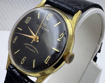 May include: A vintage Lucerna wristwatch with a round gold-tone case and a black dial. The dial features gold-colored numerals at 12, 2, 4, 6, 8, and 10, gold hands, and the text Lucerna, Unbreakable Hairspring, and Antimagnetic. A black leather strap.