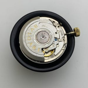 May include: A watch movement with the brand name "BVLGARI" in gold lettering. The movement is silver and gold, with intricate gears and a gold crown. It is set in a black circular holder.