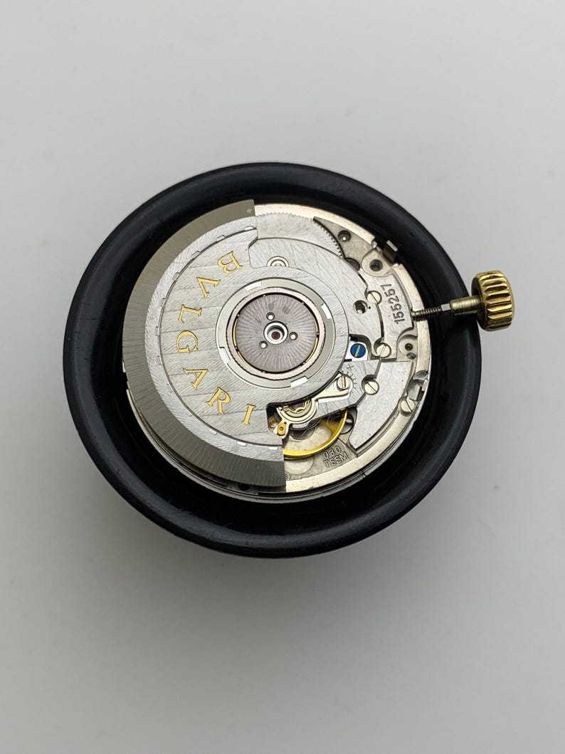 May include: A watch movement with the brand name "BVLGARI" in gold lettering. The movement is silver and gold, with intricate gears and a gold crown. It is set in a black circular holder.