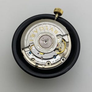 May include: A close-up of a watch movement with the brand name "BVLGARI" in gold lettering. The watch mechanism is silver with gold and blue accents, and a gold crown. The movement is set in a black holder.