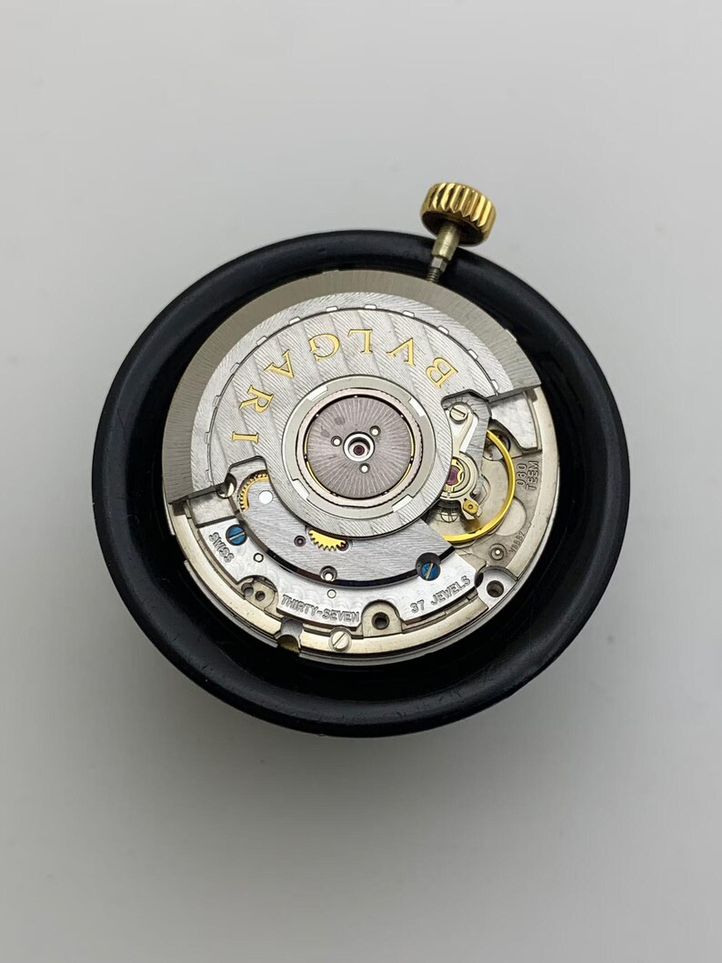 May include: A close-up of a watch movement with the brand name "BVLGARI" in gold lettering. The watch mechanism is silver with gold and blue accents, and a gold crown. The movement is set in a black holder.