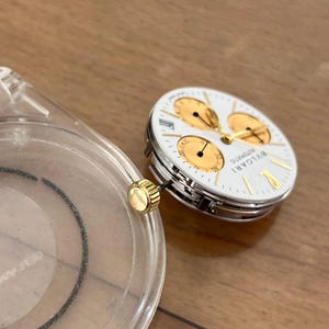 May include: A disassembled luxury watch with a white face, gold accents, and three sub-dials. The watch face is labeled "BVLGARI AUTOMATIC". A clear plastic watch case and a gold crown are also visible.