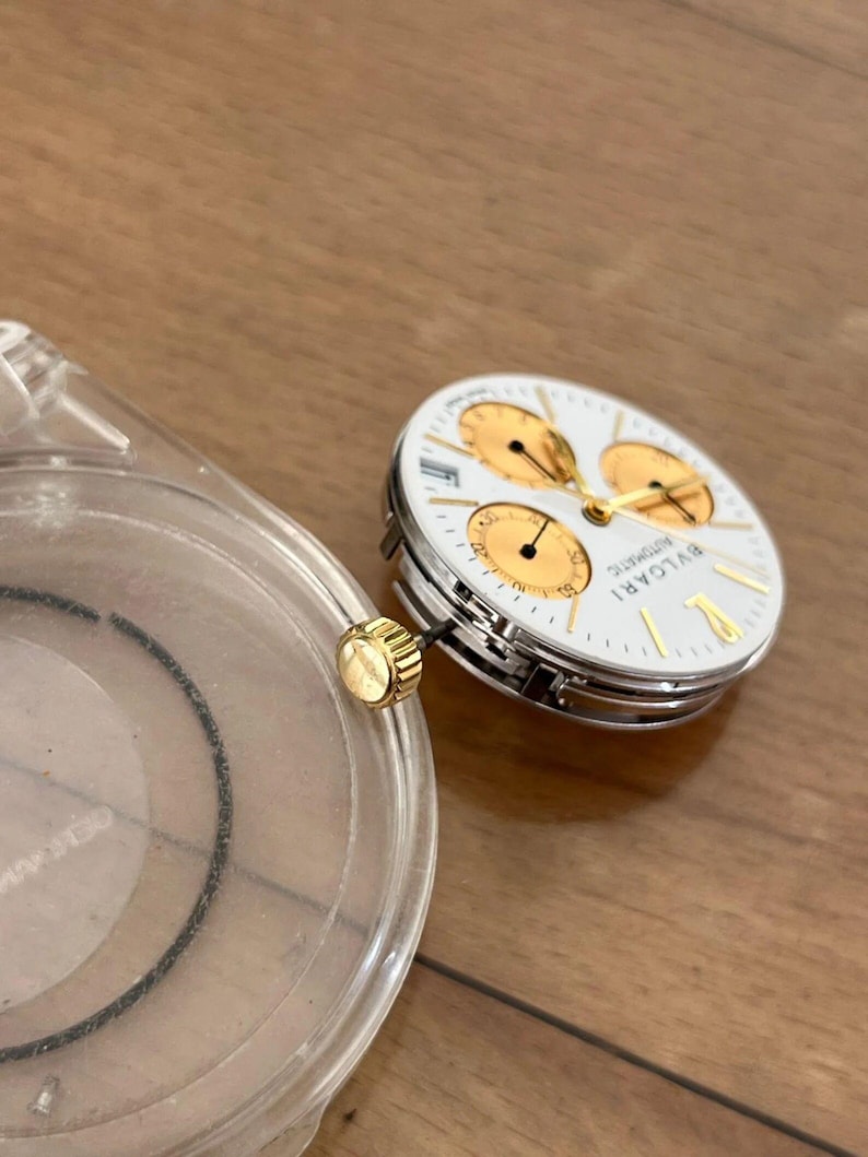 May include: A disassembled luxury watch with a white face, gold accents, and three sub-dials. The watch face is labeled "BVLGARI AUTOMATIC". A clear plastic watch case and a gold crown are also visible.