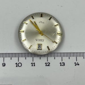 Puede incluir: Una esfera de reloj plateada con manecillas y marcadores dorados. La esfera del reloj tiene el texto "oTha", "AUTOMATIC 25 JEWELS" y una pantalla de fecha que muestra "25". Una regla está debajo.